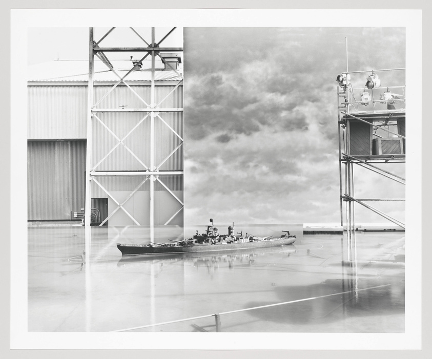 A black and white photograph showing a juxtaposition of two scenes: on the left, a large industrial building with a reflective floor, and on the right, a cloudy sky with the superimposed image of a scaffolded structure and a warship on the reflective surface.