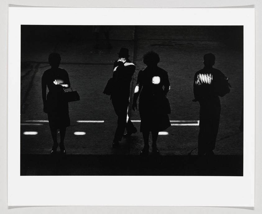 Black and white photograph depicting the silhouettes of four individuals standing on a street, with the sunlight casting their shadows on the ground and creating bright spots on their torsos. The contrast between light and shadow creates a dramatic and artistic effect.