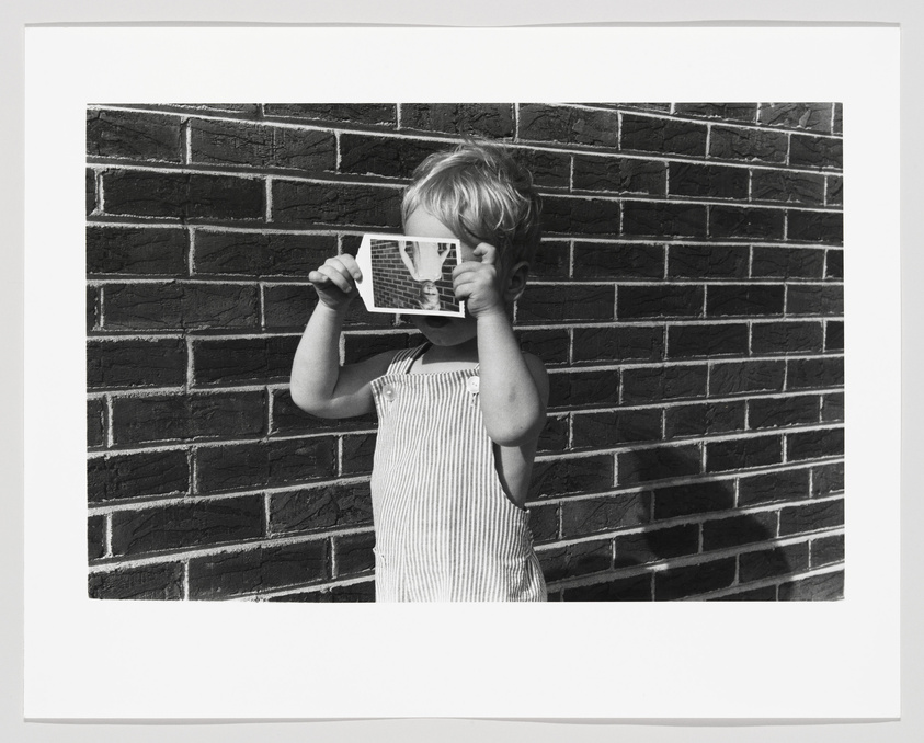 A black and white photograph of a young child standing in front of a brick wall, holding a small photograph in front of their face, aligning it with their own head to create a playful illusion. The child is wearing a striped sleeveless outfit.