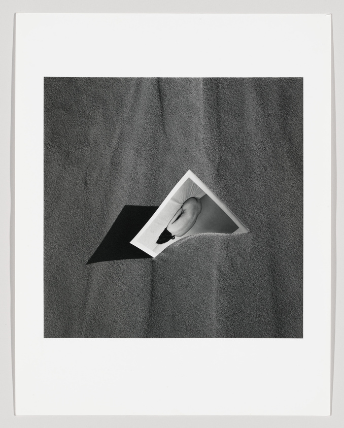 A black and white photograph featuring a hand holding a smaller photograph of the same hand, creating a visual illusion of the hand extending from the picture within a picture. The background is a textured surface, and the photo has a white border.