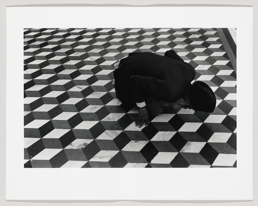 A person in dark clothing and a head covering is bowing down on a black and white checkered floor, with their hands placed on the ground in front of them. The image is in black and white, emphasizing the contrast of the patterned floor.