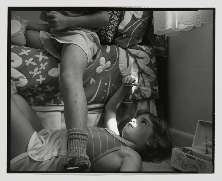 A black and white photo capturing a candid moment of a child lying on their back on the floor, gazing upward with a reflective expression. The child is surrounded by various objects, including a music box and pillows, with another person's legs partially visible in the frame. The scene is indoors, with a hint of a window treatment in the upper right corner, suggesting a cozy domestic setting.