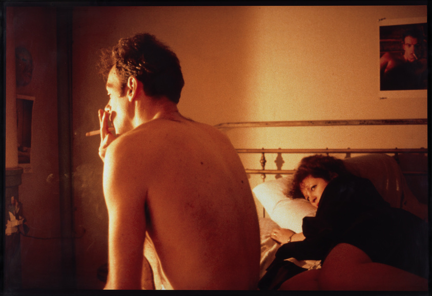 A man with his back to the camera is smoking a cigarette while a woman lies on a bed looking at him. The room is warmly lit, and there's a poster on the wall. The atmosphere appears intimate and contemplative.