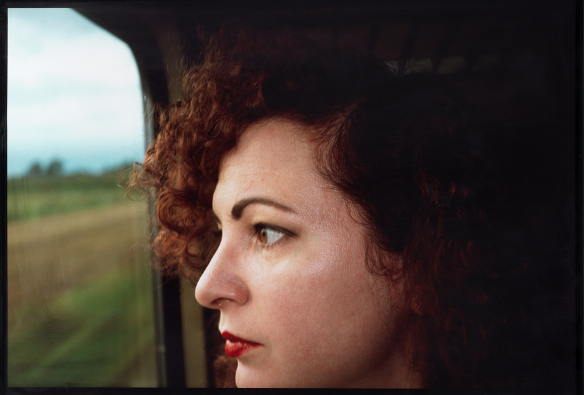 A close-up profile view of a woman with curly red hair and red lipstick looking out of a window, with a blurred landscape in the background.
