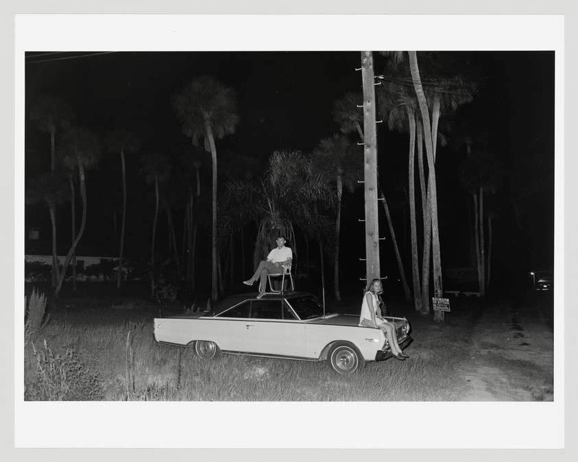 A black and white photograph depicting two individuals with a classic car at night. One person is seated on the roof of the car, while the other is sitting on the hood, both facing the camera. Tall palm trees and a utility pole are visible in the background, creating a scene that suggests a warm, tropical location. The lighting suggests the photo was taken in the evening or at night.