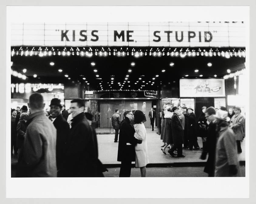 A black and white photograph of a bustling movie theater entrance with the marquee reading "KISS ME, STUPID" in large letters. People are seen mingling and walking around the entrance, with some in the foreground appearing blurred due to motion, suggesting the liveliness of the scene. The theater's facade is adorned with a string of illuminated bulbs, adding to the classic cinematic ambiance.