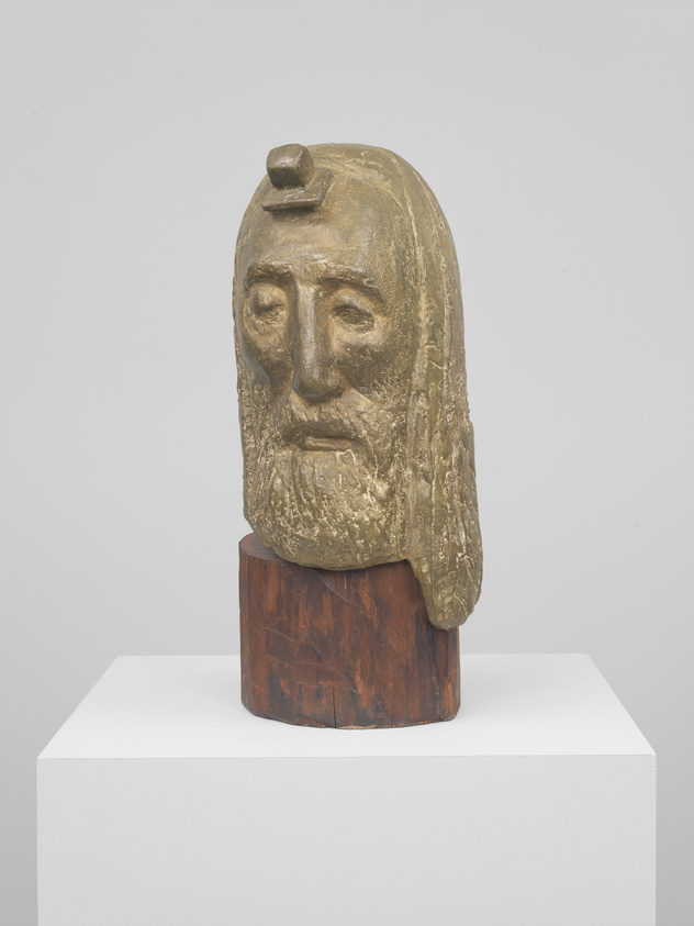 A bronze sculpture of a bearded man's head with a solemn expression, mounted on a wooden base, displayed on a white pedestal against a light background.
