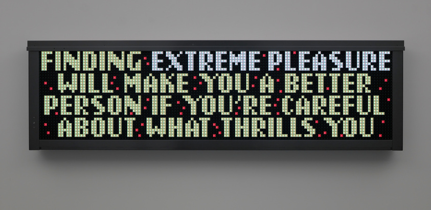 A digital display board with a message in bright white and green lights against a black background that reads "FINDING EXTREME PLEASURE WILL MAKE YOU A BETTER PERSON IF YOU'RE CAREFUL ABOUT WHAT THRILLS YOU." Red lights punctuate the display at various points.