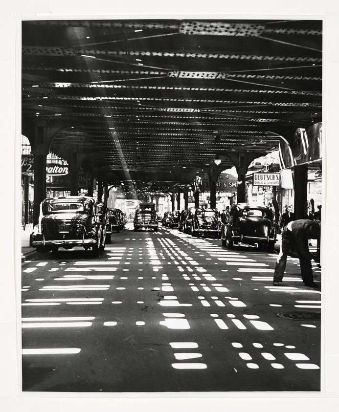 Black and white photograph depicting a bustling city street under an elevated train track, with vintage cars and pedestrians. Sunlight filters through the overhead structure, casting a pattern of light and shadows on the vehicles and roadway. Signs for businesses, including one that reads "Deutsch's," are visible along the sidewalk.