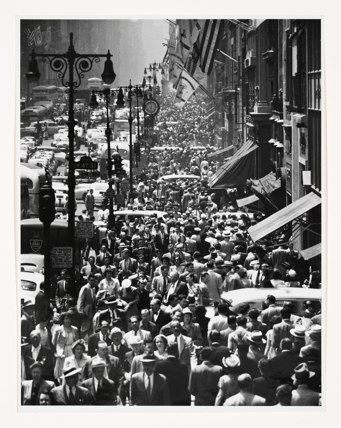 A bustling city street scene in black and white, crowded with pedestrians and vintage cars. People are dressed in mid-20th-century attire, suggesting a historical setting. The street is lined with buildings adorned with flags, and ornate street lamps are visible. The density of the crowd and the number of vehicles suggest a busy urban environment, possibly during a special event or rush hour.