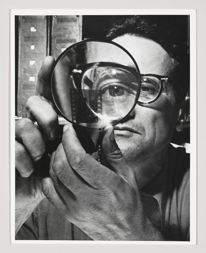 A black and white photo of a man holding a large magnifying glass up to his eye, which magnifies the eye and the frames of his glasses. Behind him are out-of-focus images that appear to be film negatives.