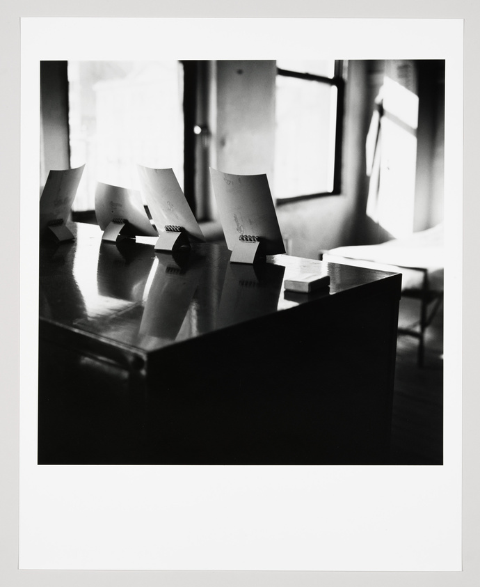 Black and white photograph of several sheets of paper with binder clips attached, standing upright on a wooden surface, with light streaming in from a window in the background creating a high-contrast scene.