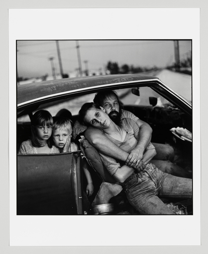 Black and white photograph of a family with two adults and two children sitting inside an old car, looking directly at the camera with solemn expressions. The background shows a blurred landscape with utility poles.