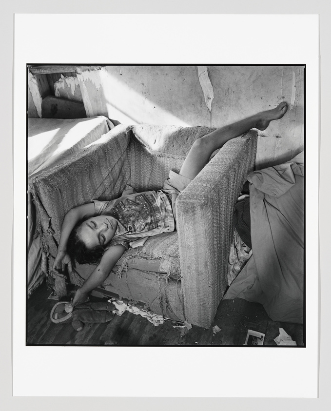 Black and white photograph of a person lying on a worn-out couch with exposed stuffing, in a relaxed pose with one arm draped over their forehead and one leg propped up against the back of the couch. Sunlight filters into the room, casting shadows on the wall and couch. The scene suggests a moment of rest in a dilapidated interior setting.