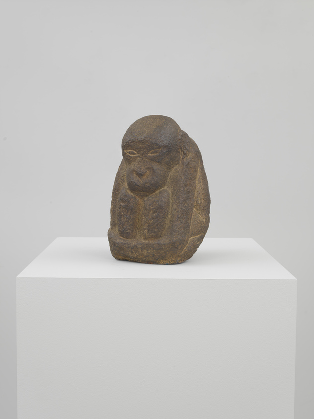 A stone sculpture of a seated baboon with pronounced facial features, displayed on a white pedestal against a plain background.