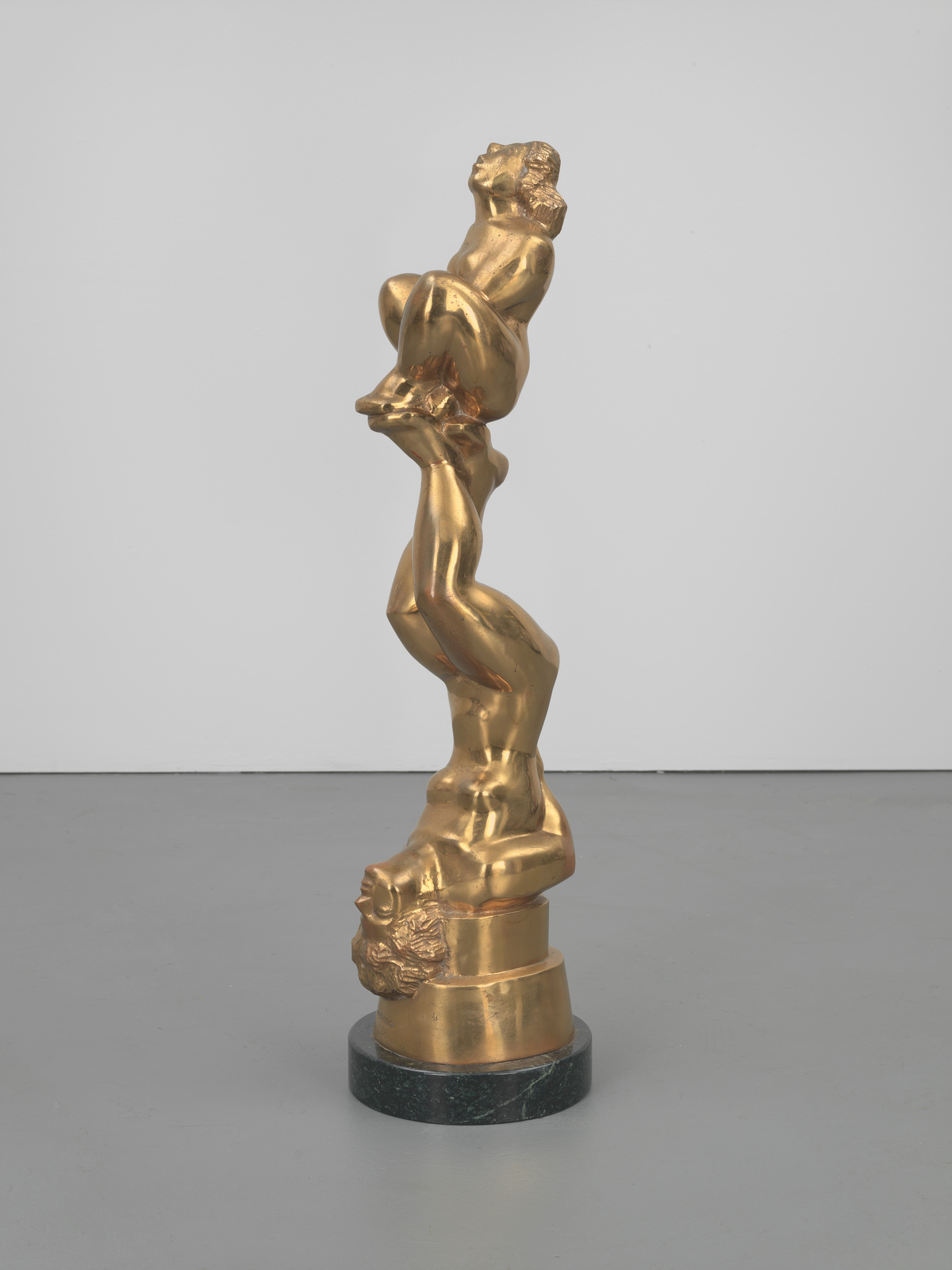 A golden sculpture of a twisted human figure composed of several bodies stacked vertically, each contorted in different postures, displayed on a dark circular base.