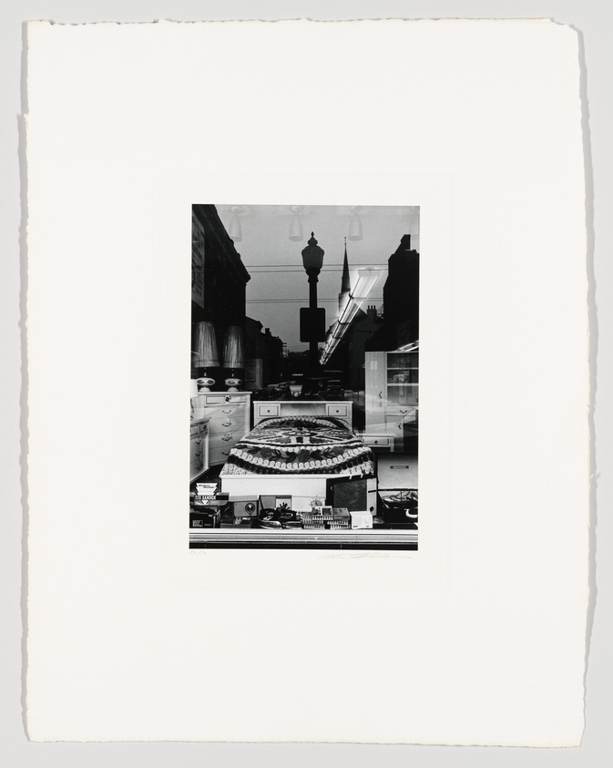 Black and white photograph depicting a street scene reflected in a shop window, showcasing a variety of small items on display, with buildings and street lamps visible in the reflection. The image is centered on a textured white background.