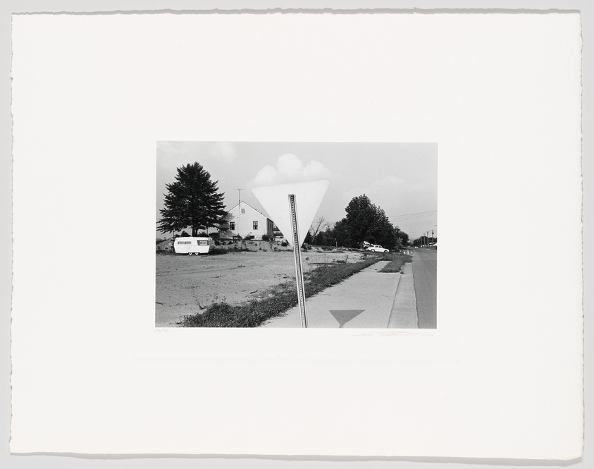 Black and white photograph depicting a suburban landscape with a house and trees in the background, a yield road sign in the foreground to the left, and a sidewalk running parallel to a street on the right. The image has a white border, suggesting it is a print.