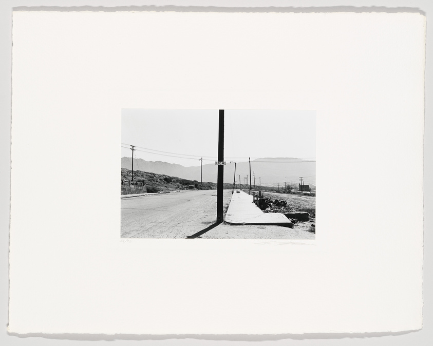 Black and white photograph depicting a desolate street scene with a utility pole centered in the middle of the frame, dividing the image into two symmetrical halves. A street sign reads "DEAD END" on the right side, with arid hills in the background and a clear sky above. The photo is mounted on a white border.