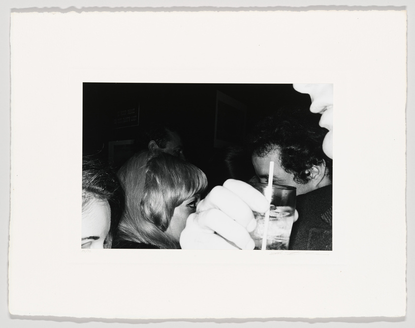 A black and white photograph capturing a candid moment among a group of people, with a partial view of a person's face in the foreground on the right, someone holding a glass with a straw in the center, and various other individuals in the background, suggesting a social gathering or event. The image has a vintage feel, indicated by the white border typical of old photographs.