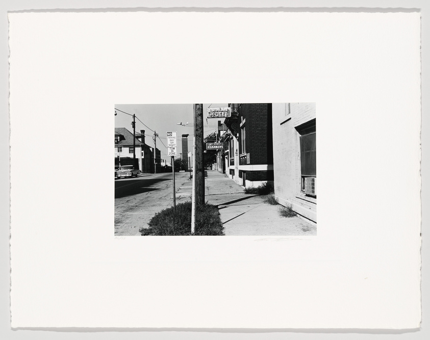 Black and white photograph depicting a mid-20th-century street scene with vintage cars parked along the curb, various storefronts including a hotel and a cleaners, and a "NO PARKING" sign prominently displayed in the foreground. The image has a white border typical of old photographs.