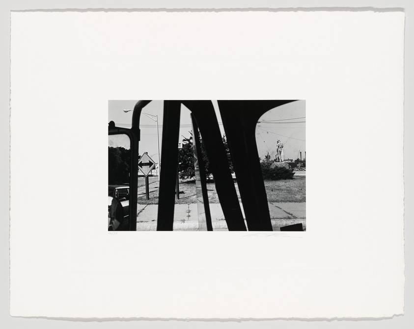 Black and white photograph framed by the silhouette of a car window, showing a street scene with a car, road signs, and a statue in the distance, all under a clear sky. The image has a white border with rough edges, suggesting it is a print.
