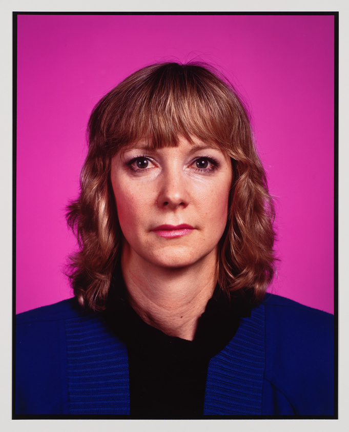 Woman with shoulder-length blonde hair and blue jacket looks straight ahead against a bright pink background.