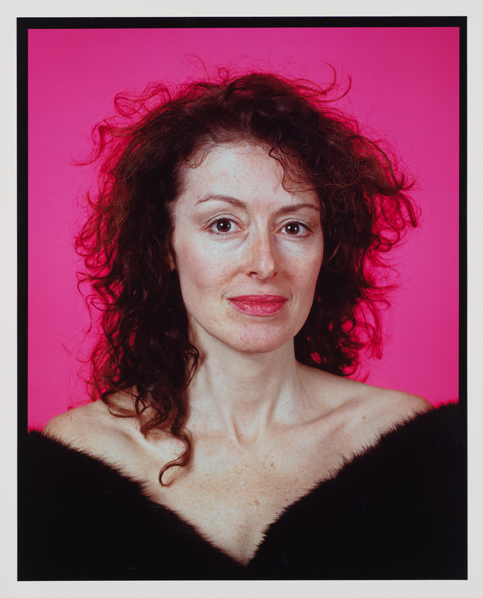 A portrait of a woman with curly auburn hair against a vibrant pink background. She is wearing a black off-the-shoulder top and has a subtle smile. Her makeup is natural with a touch of pink lipstick.