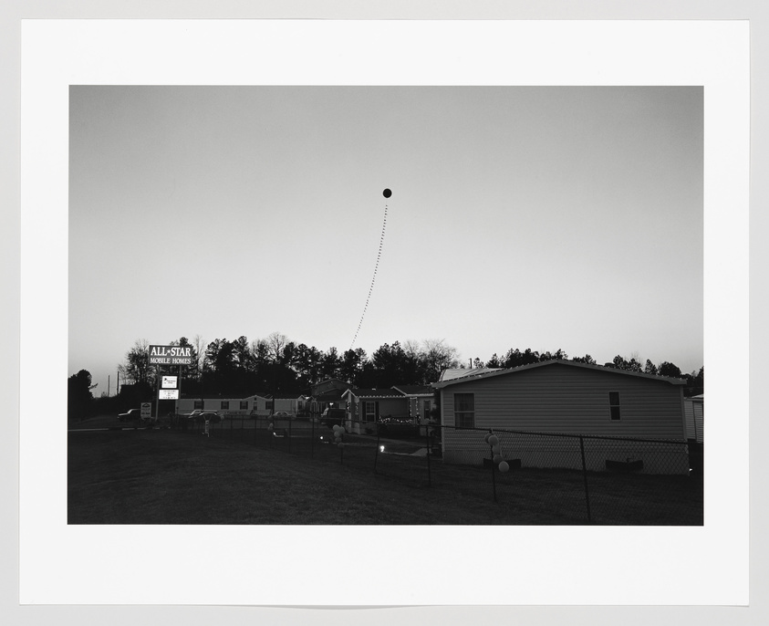 Black and white photo of a mobile home park with a dotted line leading to a balloon in the sky.