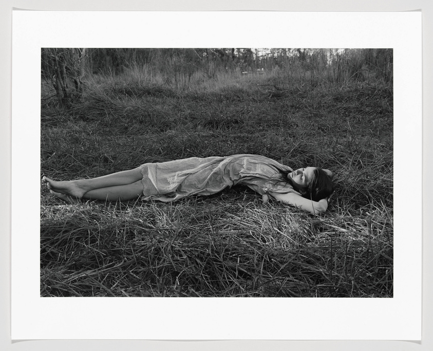 A black and white photograph of a woman lying on her side in a field of tall grass, wearing a light, flowing dress. Her eyes are closed, and her arms are folded under her head as if she is resting or sleeping. The background shows a hint of trees and a subdued sky, suggesting a tranquil, natural setting. The image is framed with a white border.