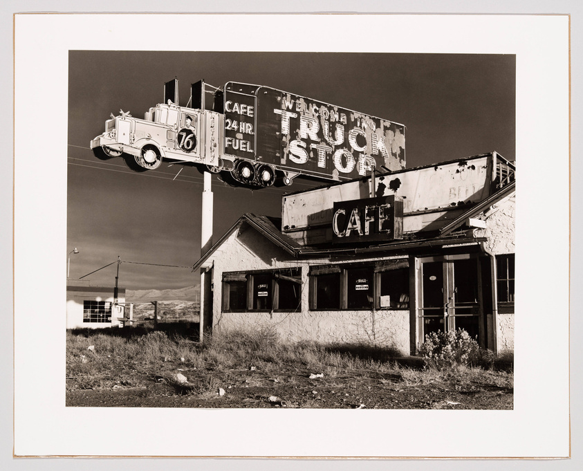 A black and white photograph depicting an old, dilapidated truck stop with a large, vintage billboard above it. The billboard features a three-dimensional truck and trailer model and advertises a 24-hour cafe and fuel. The words "WELCOME TRUCK STOP" are visible on the sign, which towers over the abandoned building below. The surrounding landscape is barren and the sky is clear.