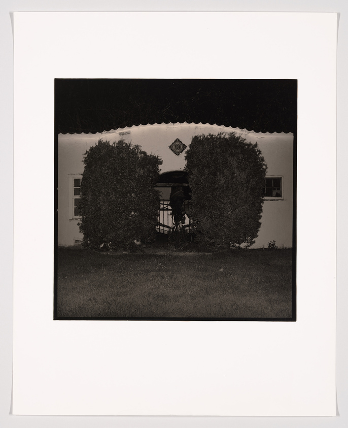 A black and white photograph depicting the front of a house with a symmetrical design, featuring two large bushes trimmed into rounded shapes on either side of a central pathway leading to a porch with a decorative diamond-shaped motif above the entrance. The image is framed with a wide white border.