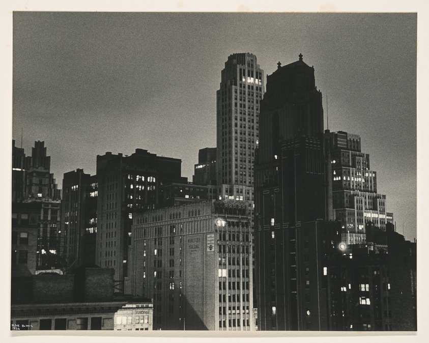 A black and white photograph capturing the silhouette of a city skyline at dusk, with the outlines of various high-rise buildings against a fading light sky. Some windows are illuminated, adding contrast to the darkened structures. The architecture suggests an urban setting from an earlier era, possibly mid-20th century.