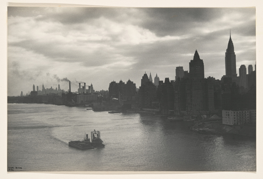 Black and white photograph of a vintage New York City skyline with the Empire State Building prominent against a backdrop of clouds. A river in the foreground reflects light, and a tugboat is seen moving on the water. Smokestacks emit smoke on the left, contributing to the industrial atmosphere of the scene. The photo is marked with "Ilse Bing 1936" on the bottom left corner.