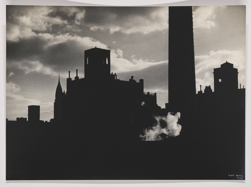 Silhouetted cityscape against a cloudy sky with a prominent smokestack on the right and a puff of smoke on the lower left, captured in black and white. The photo is signed "Ilse Bing 1936" in the bottom right corner.
