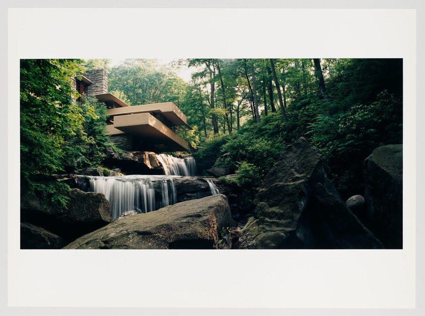 A modern architectural house with multiple cantilevered levels over a natural waterfall surrounded by a lush forest. Rocks and foliage are visible in the foreground.