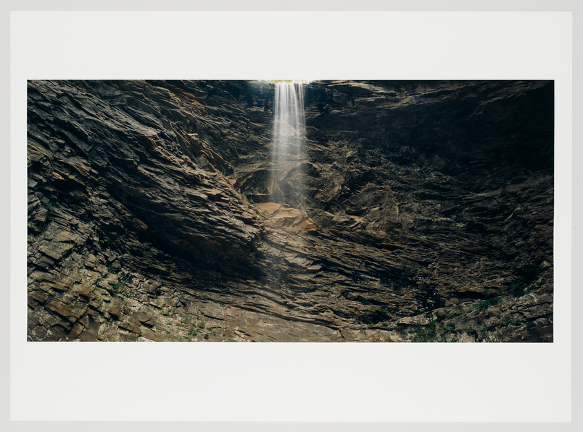 A slender waterfall cascades down a rugged cliff face, with sunlight filtering through the mist at the top. The rocky surface surrounding the waterfall is layered and textured, with sparse vegetation clinging to the crevices.