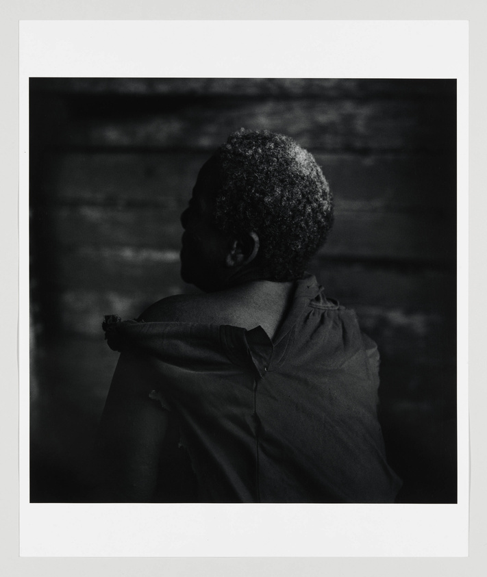 Black and white photograph of an elderly person with short, curly gray hair, viewed from behind and looking to the side. The individual is wearing a worn-out, sleeveless shirt with a torn section near the shoulder. The background is dark and textured, suggesting an old wooden surface. The photo has a white border framing the image.