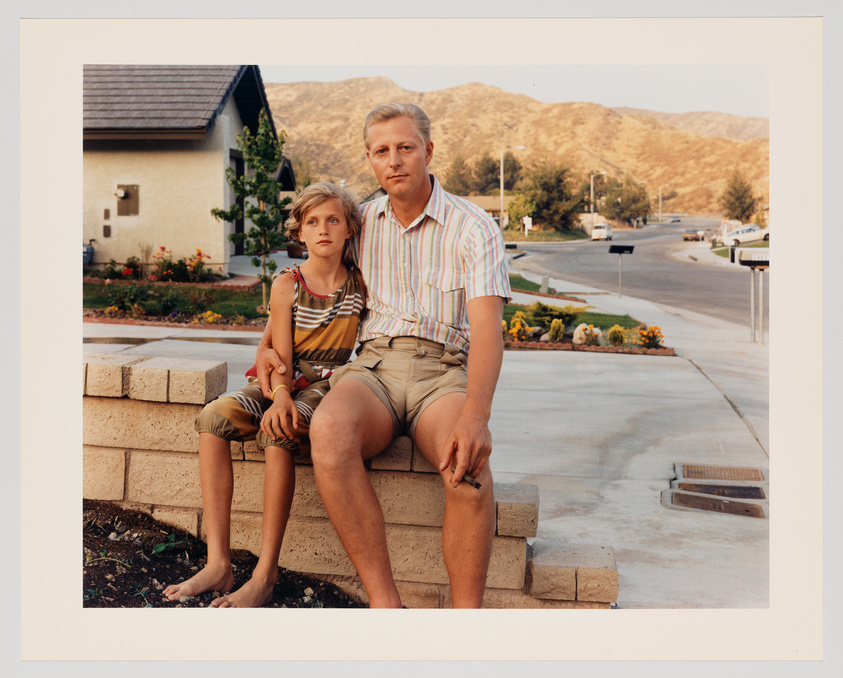 A young girl and an adult man sit side by side on a low brick wall in front of a house with a well-manicured lawn and a backdrop of rolling hills. Both are barefoot and dressed in summer clothes, looking directly at the camera with neutral expressions. The street behind them is quiet and residential, with a clear sky above.