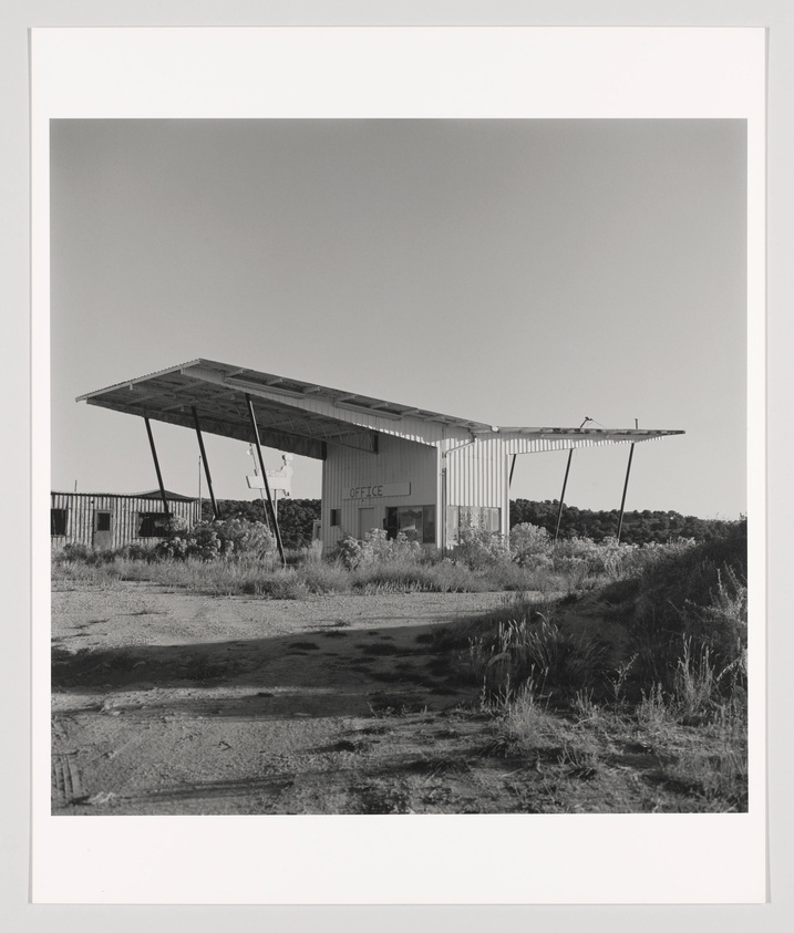 A black and white photograph of an abandoned building with a large, angled roof that extends beyond the structure's walls. The word "OFFICE" is visible on the side of the building. Overgrown vegetation and a cracked concrete path are in the foreground, with a clear sky in the background.