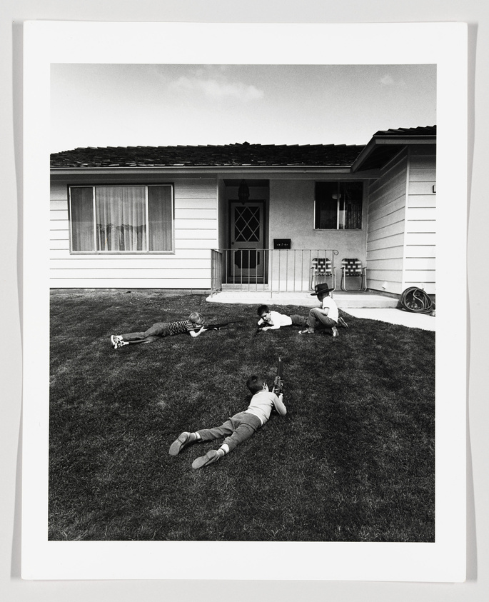 Black and white photograph depicting four children playing on a well-manicured lawn in front of a single-story suburban house. One child lies on their stomach, another sits upright, and two others are sprawled on the grass, with a toy rifle visible next to one of them. The house features a large window, a small porch with a railing, and two chairs. A bicycle lies on its side in the background.