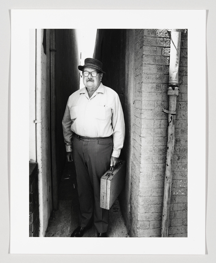 A black and white photograph of an older man standing in a narrow alleyway. He is wearing glasses, a hat, a light-colored shirt, and striped trousers. He holds a briefcase in his right hand and has a stern expression on his face. The alley walls are textured, and there's a pipe running down the wall to his right.