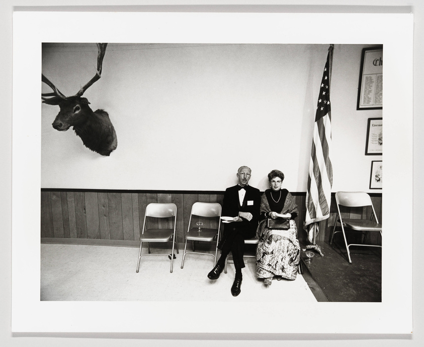 A black and white photograph depicting an elderly couple seated side by side on metal folding chairs against a plain wall, with a mounted deer head above them and an American flag to their right. The couple appears to be dressed formally, with the man wearing a suit and bow tie and the woman in a patterned dress and necklace, both looking directly at the camera. The setting suggests a community hall or similar venue.