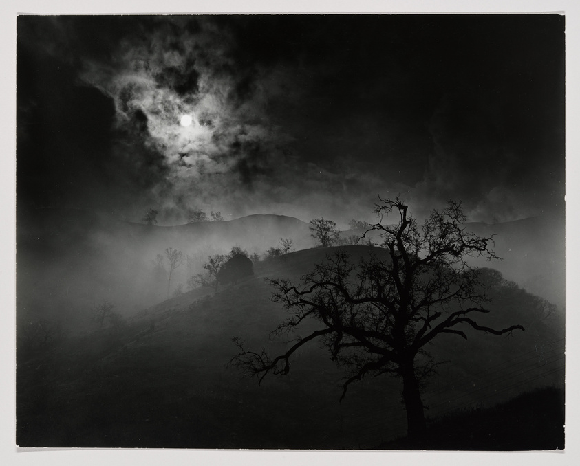 A black and white photograph depicting a dramatic night scene with a full moon partially obscured by clouds. Mist or fog blankets rolling hills, and the silhouette of a leafless tree with gnarled branches stands prominently in the foreground. The overall mood is eerie and mysterious.