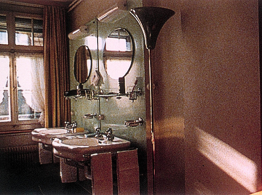 A vintage bathroom with two sinks, round mirrors, and a large window with curtains. A floor lamp stands to the right, casting a warm light.