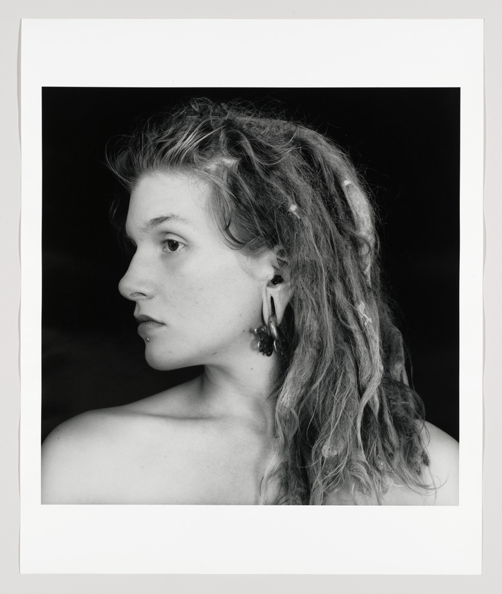 Black and white profile portrait of a woman with shoulder-length wavy hair, featuring a bare shoulder and wearing an earring. The background is dark, and the photo has a white border.
