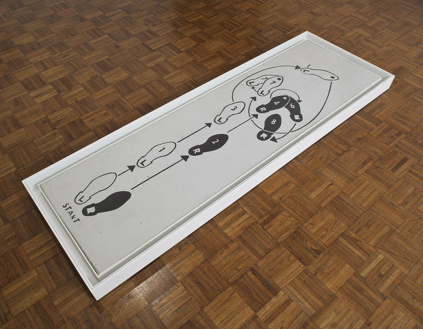 A framed piece of artwork is lying on a herringbone patterned wooden floor. The artwork features a simplistic black and white line drawing with abstract figures and shapes, including circular forms and arrows, with the word "START" at one end.