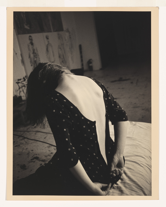 A black and white photograph of a person seated on a striped mattress, bending forward with their head down and arms reaching towards their feet. The person is wearing a polka-dotted dress, and the background features a canvas with sketches and a sparse, dimly lit room.