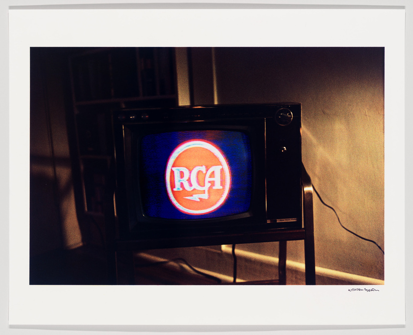 A vintage television set on a small stand displaying the RCA logo with vivid red and blue colors on its screen, situated in a dimly lit room with a bookshelf in the background.