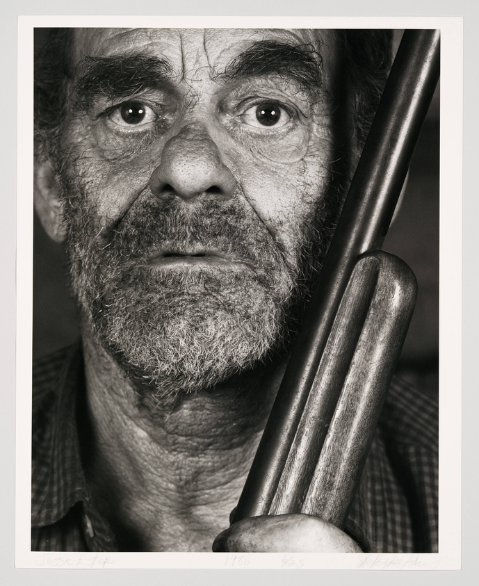 Close-up black and white portrait of an older man with a weathered face, holding a baseball bat over his shoulder. He has a beard and intense eyes, and is wearing a plaid shirt. The photograph has a signature and edition number on the bottom.